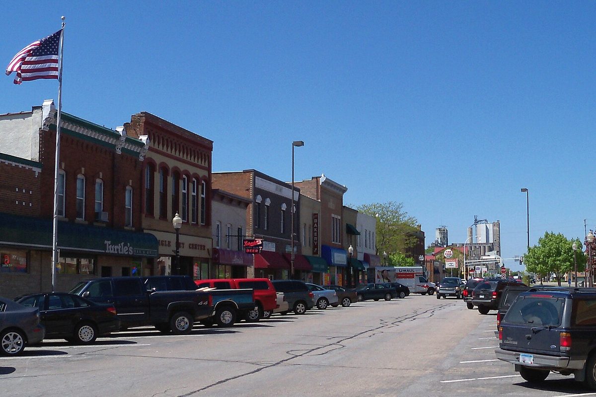 Downtown Shakopee's main street: two- and three-story brick storefronts with awnings, an American flag on a pole and cars parked along the curb