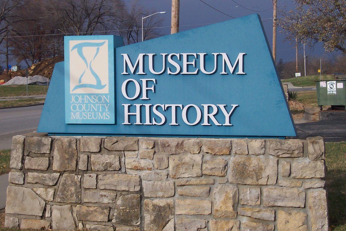 The blue Museum of History sign for Johnson County Museums on a stone base in Shawnee, with bare trees and a road behind it