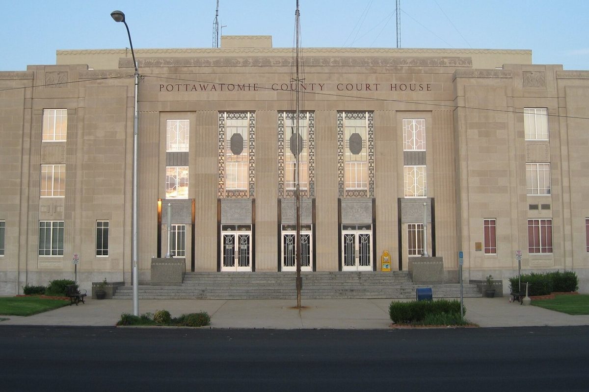 The pale stone front of the Pottawatomie County Court House in Shawnee, its name carved above three sets of tall doors, with steps and a lamppost in front