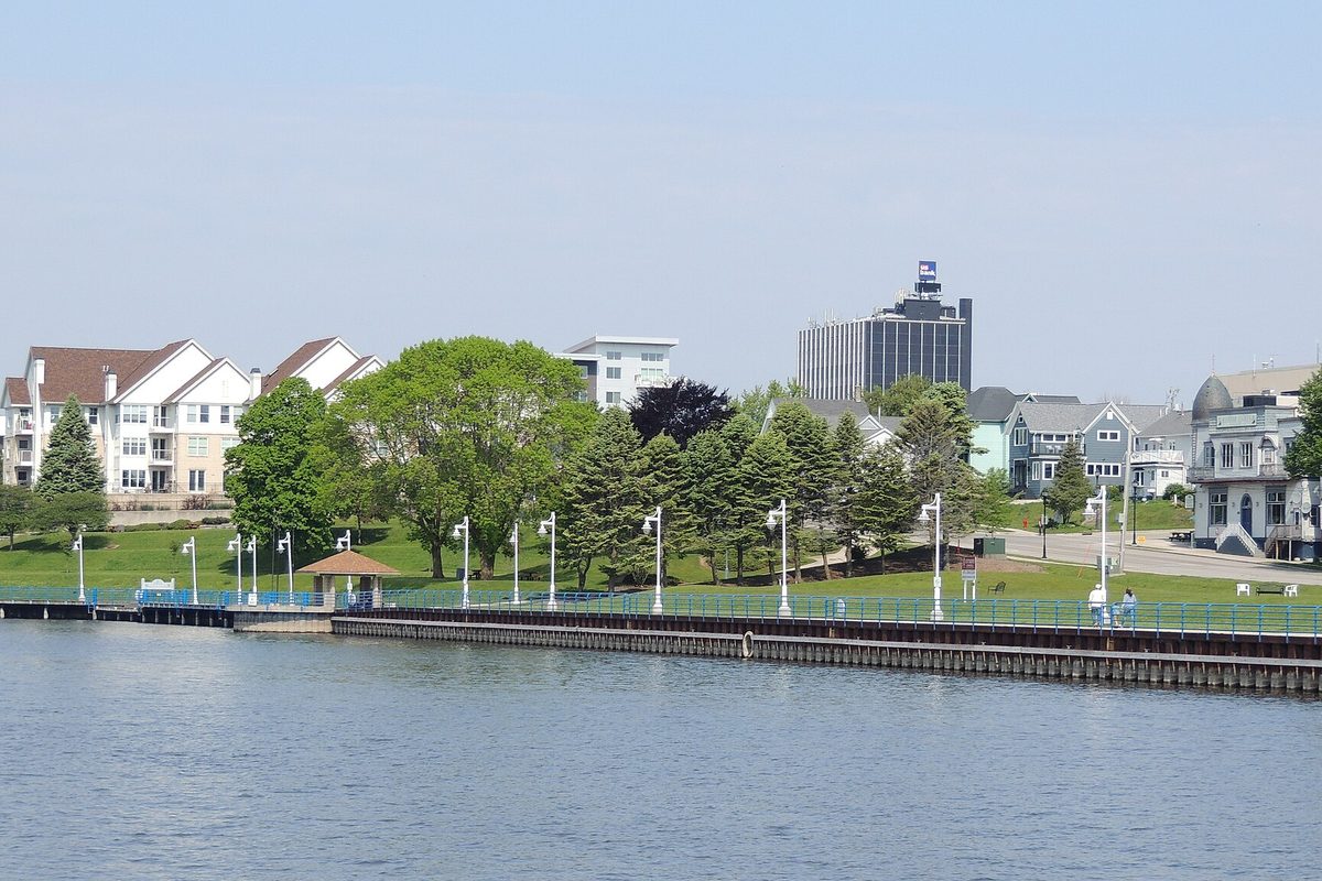 The Sheboygan riverfront with a boardwalk, lamp posts, a small gazebo, green lawns and houses and a tall office building above the water