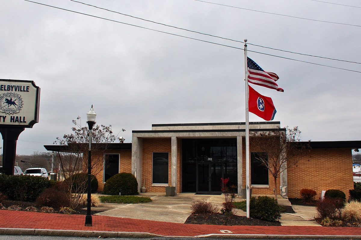 Shelbyville City Hall, a flat-roofed brick building with the American and Tennessee flags out front and the city hall sign at the left