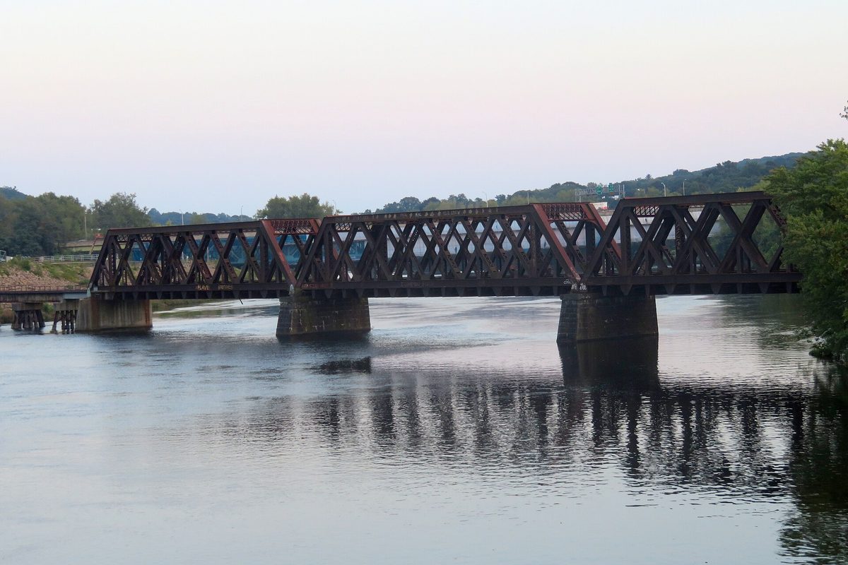 A rust-brown steel truss railroad bridge on concrete piers crossing the calm Housatonic River at dusk, with wooded hills behind