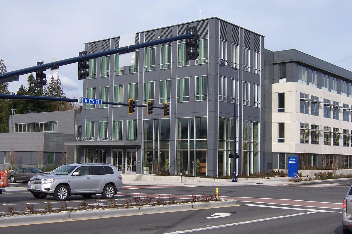 Shoreline City Hall, a modern gray building with tall green-tinted windows at the corner of Midvale Avenue and North 175th Street, with traffic lights and a silver SUV in front