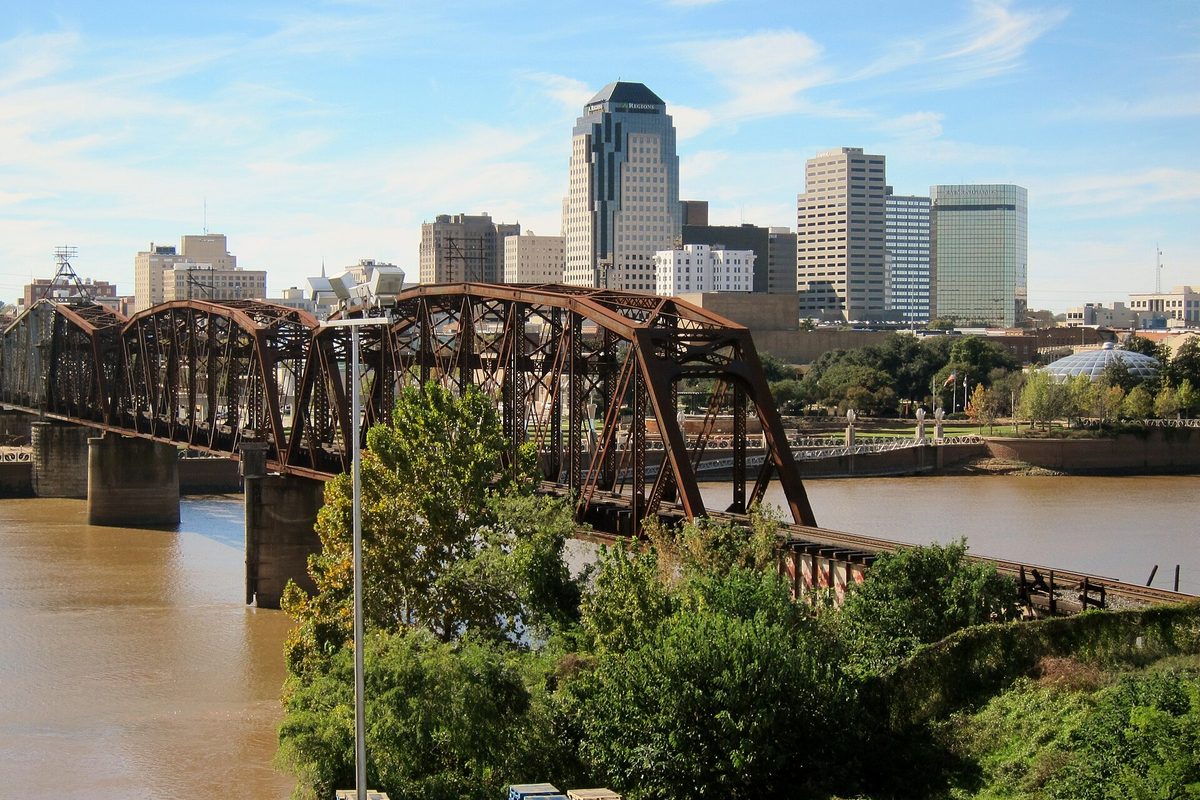 A rusted steel truss bridge crossing the brown Red River, with downtown Shreveport's towers rising behind the trees