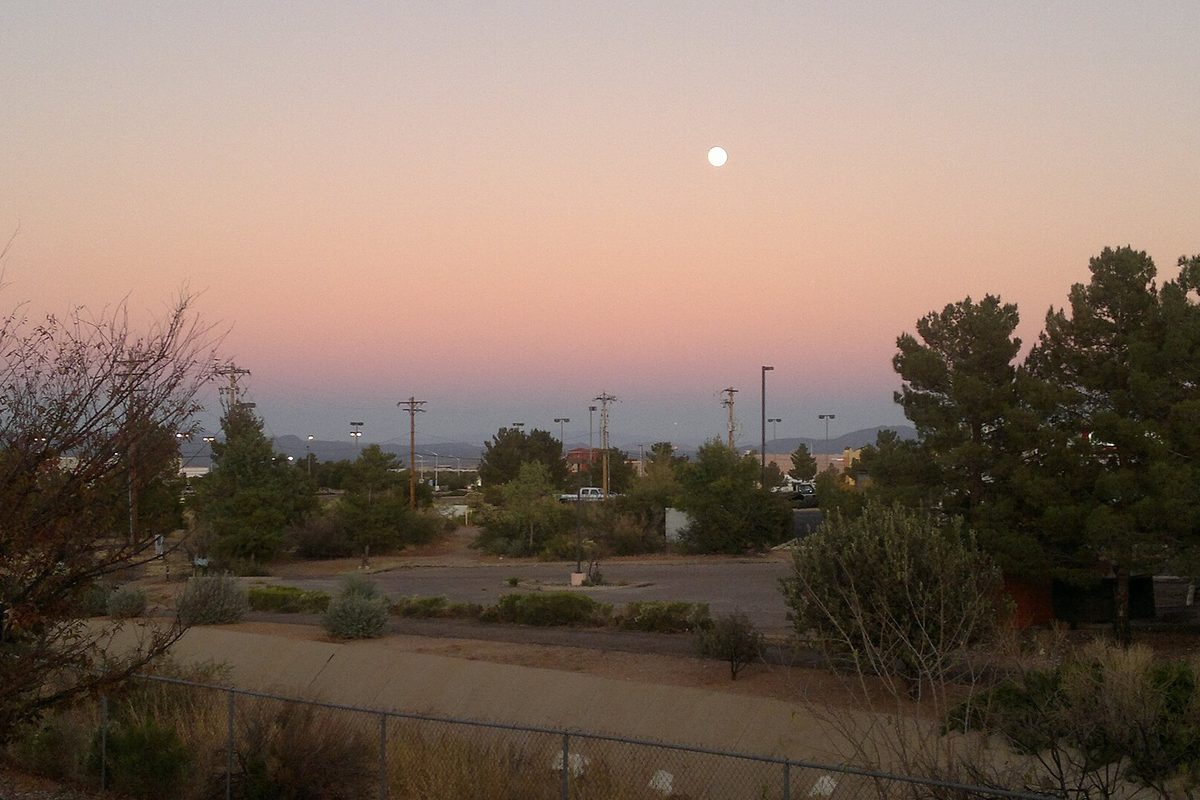 A pink and blue dusk sky with a full moon over Sierra Vista, above desert trees, power poles, a parking lot and distant hills