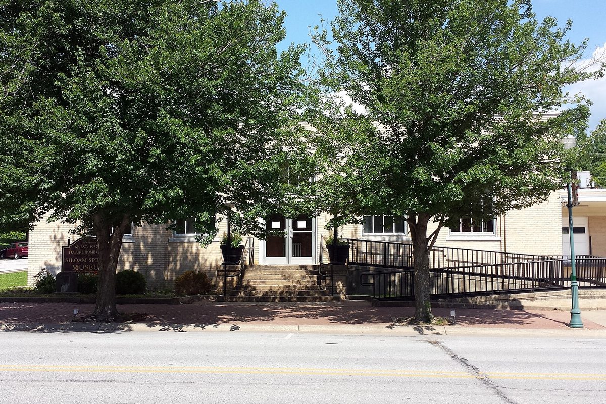 The old post office in downtown Siloam Springs, now the Siloam Springs Museum, a pale brick building with front steps behind two leafy trees