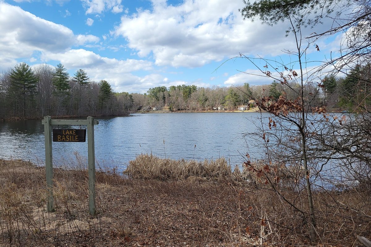Lake Basile in Simsbury on a bright spring day, with a small wooden sign on posts in dry grass at the water's edge and bare trees along the far shore