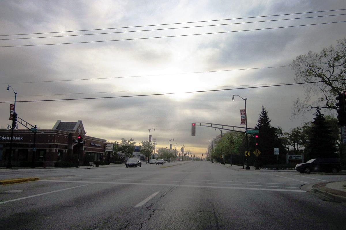 A wide, empty Skokie street at an intersection under a cloudy sky, with the brick Edens Bank building on the left corner