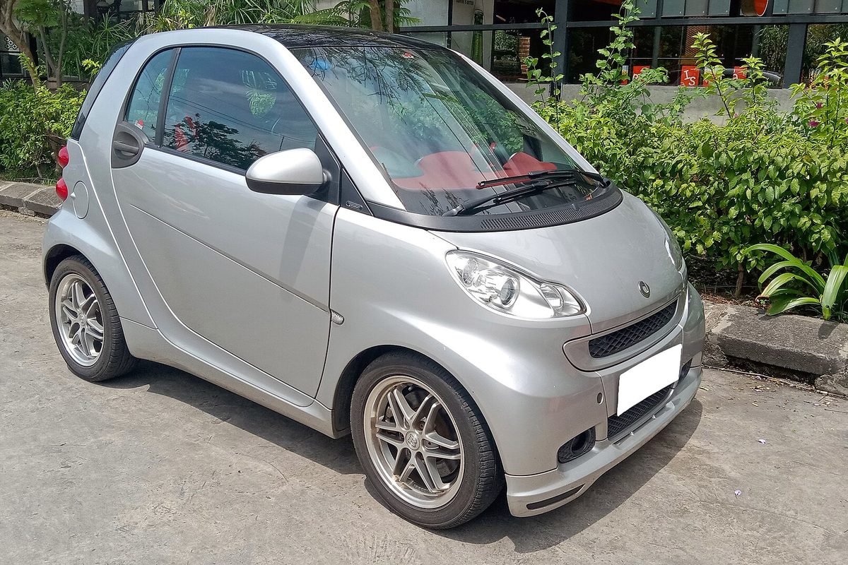 A silver smart fortwo coupe with multi-spoke alloy wheels and a red interior, parked on concrete beside a row of shrubs
