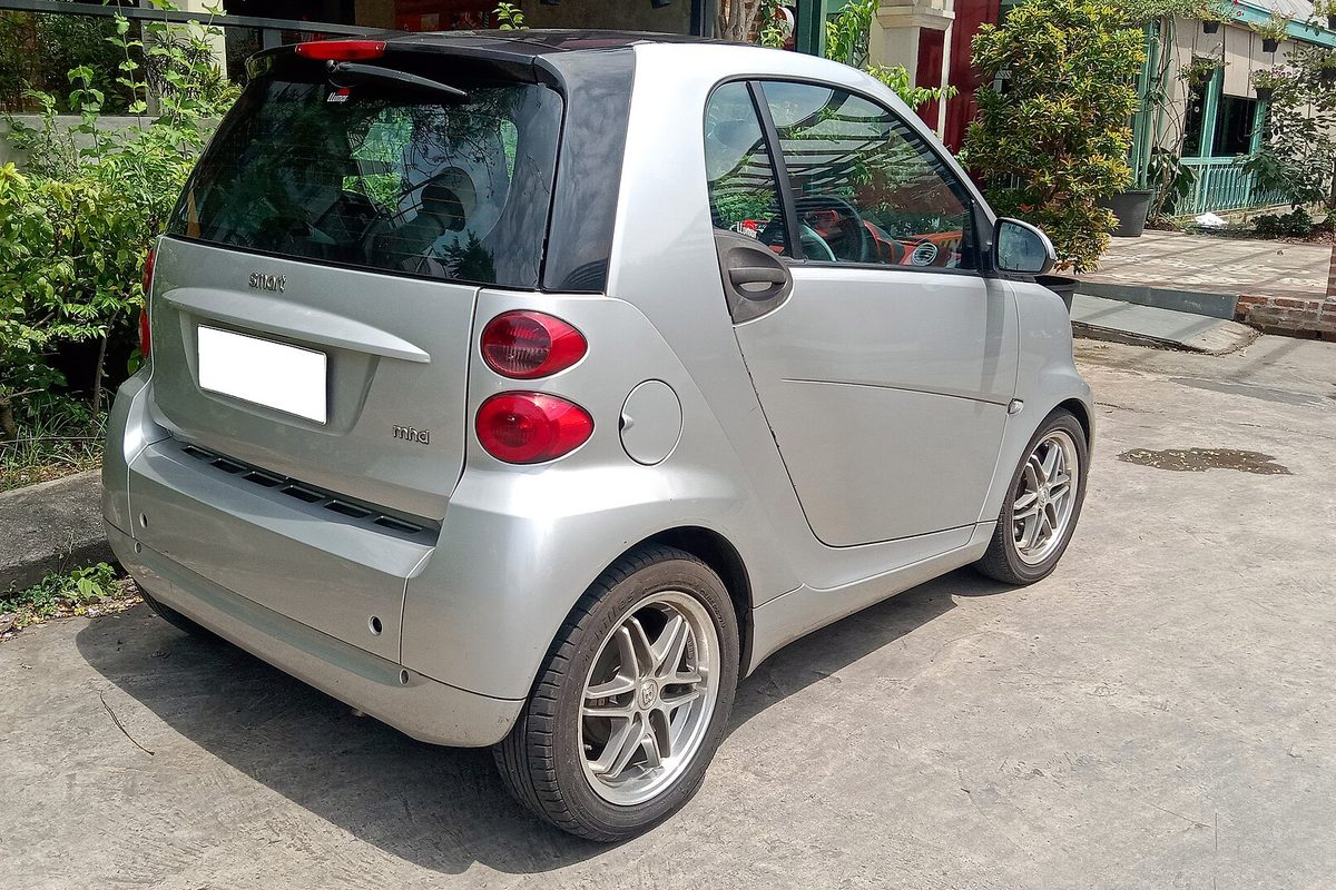 A silver smart fortwo coupe seen from the right rear, parked on a concrete drive beside a hedge