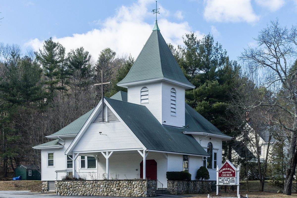 The white wooden Georgiaville Baptist Church in Smithfield, with a green-roofed steeple and a fieldstone wall in front, pine trees behind