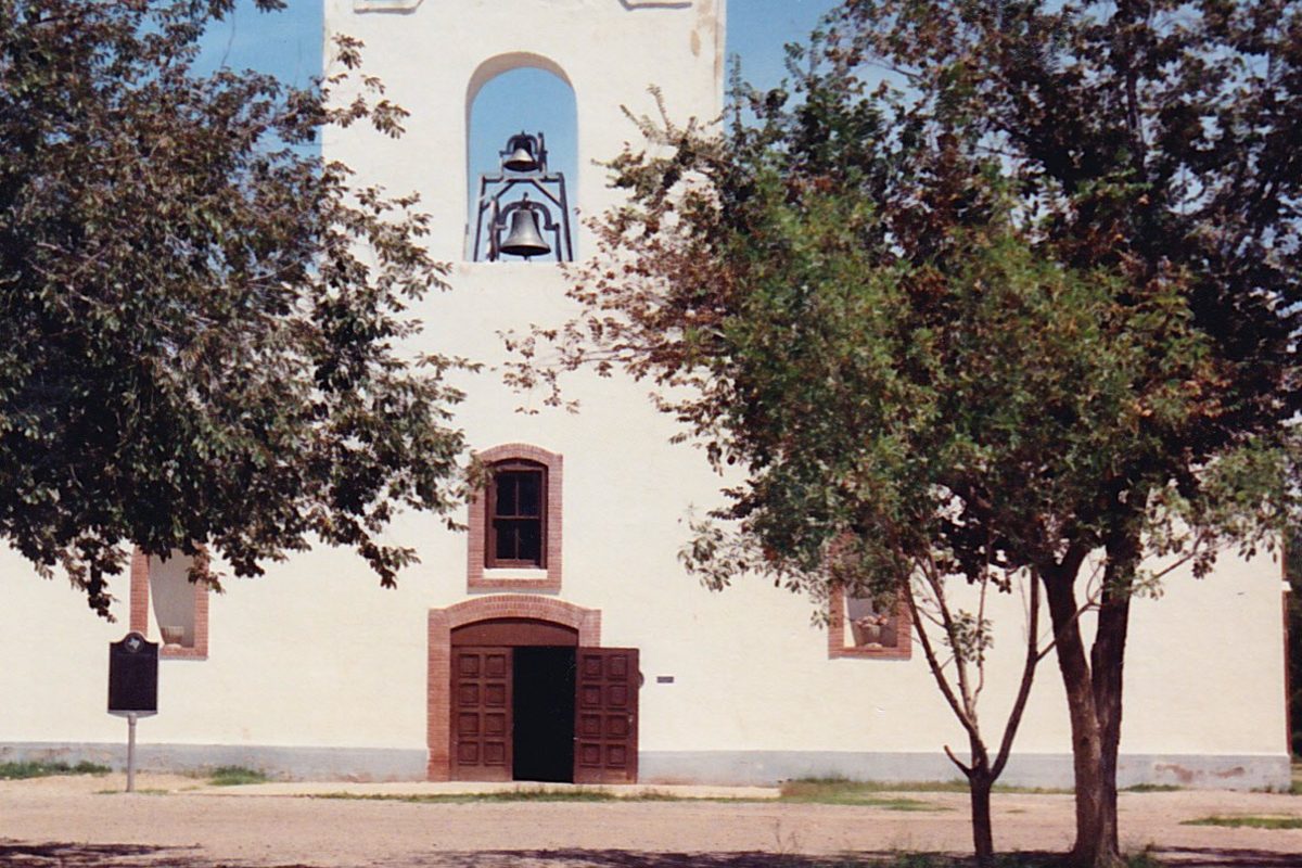 A whitewashed adobe church with a bell hanging in its open tower and a brick-framed wooden door, half hidden by two shade trees in Socorro, Texas
