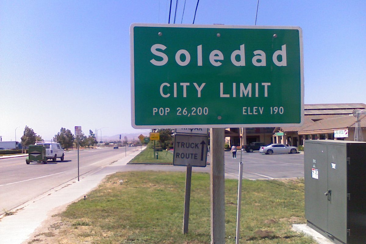 Green Soledad City Limit sign with population and elevation figures, beside a truck route sign on a sunny roadside