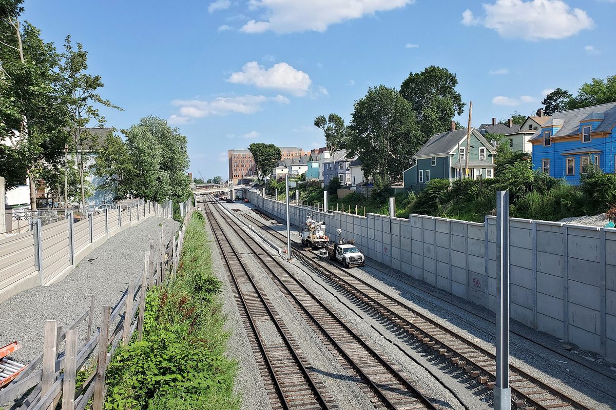 Railroad tracks in a cut beside a tall concrete retaining wall, two utility trucks on the rails and wooden houses on the hill above, Somerville