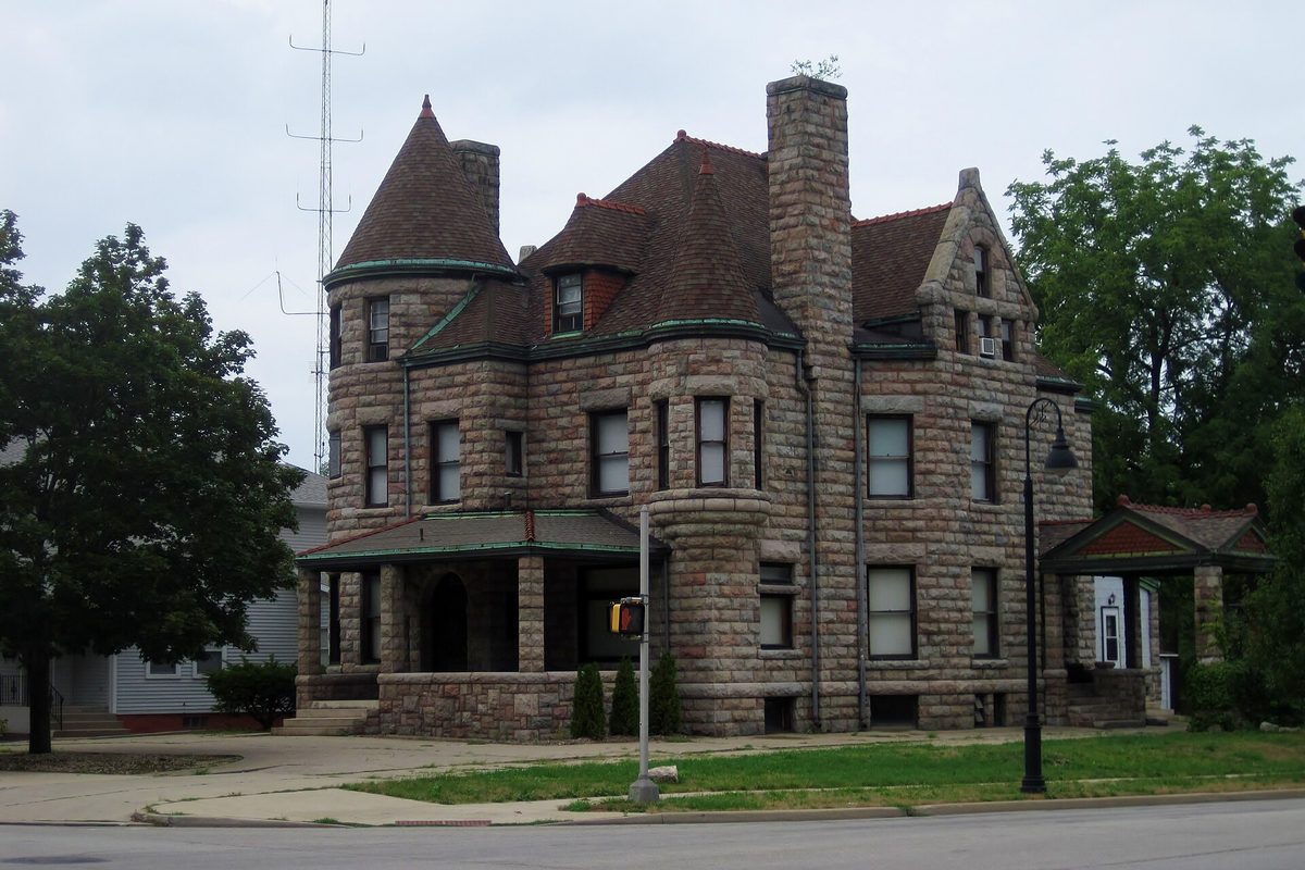 The stone Kaiser-Schmidt House in South Bend, with a round corner tower, a tall chimney and a green-trimmed porch, under a gray sky