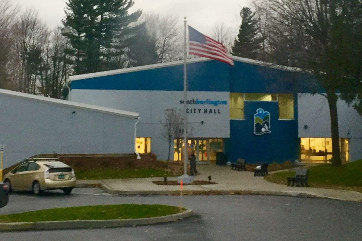 South Burlington City Hall, a blue and white building with lit windows and an American flag on a pole out front, evergreen trees behind it at dusk