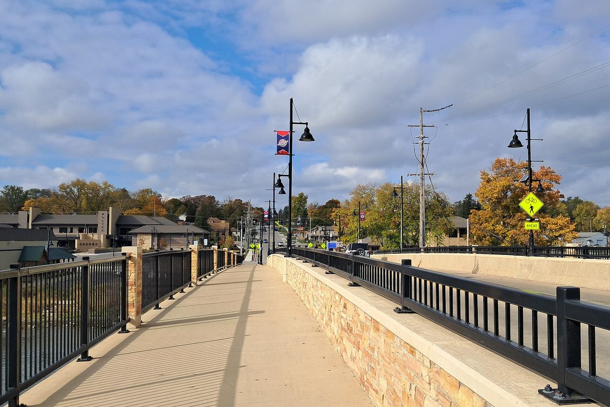 The walkway on the State Street Bridge in South Elgin, with black railings, lampposts with banners and fall trees under a cloudy sky
