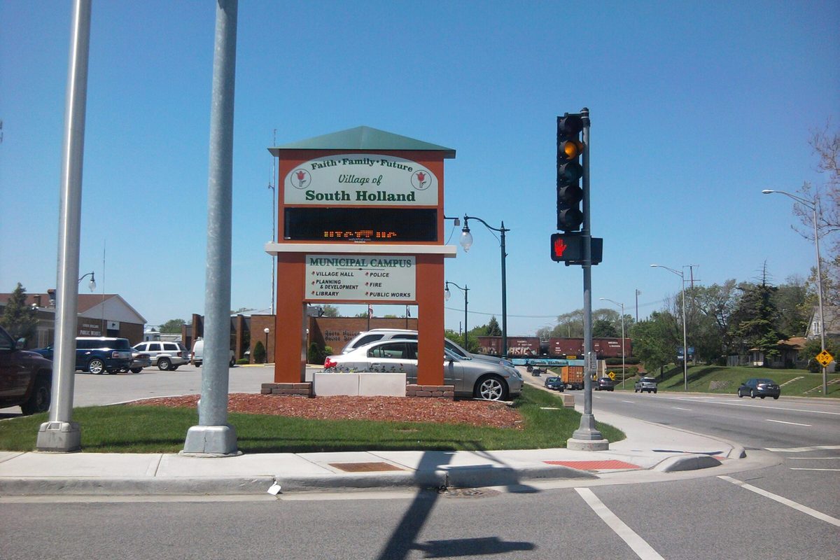 The Village of South Holland municipal campus sign at a street corner, with a traffic signal, parked cars and a clear blue sky