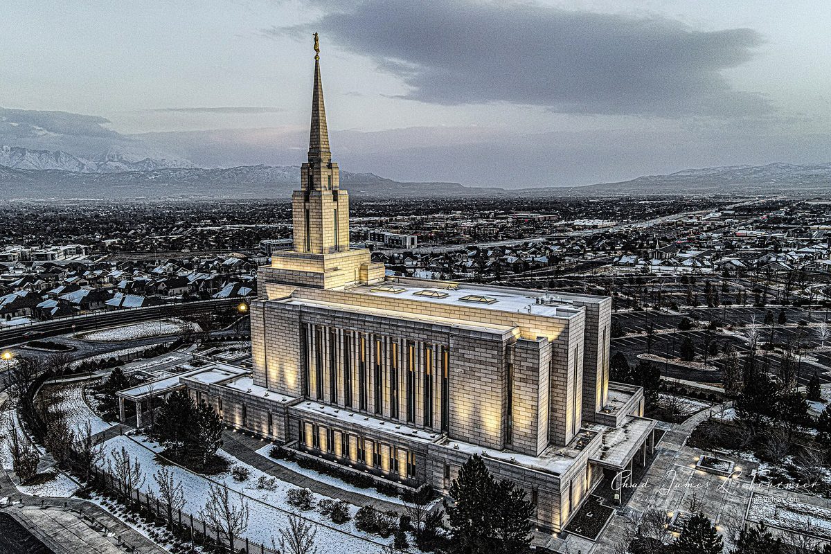 The Oquirrh Mountain Utah Temple lit up at dusk on a bluff, with snow-dusted neighborhoods and mountains stretching behind it