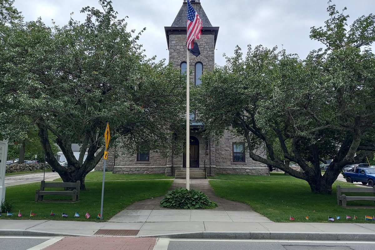 The stone South Kingstown town hall with its square tower and an American flag out front, framed by two leafy trees and a lawn with benches