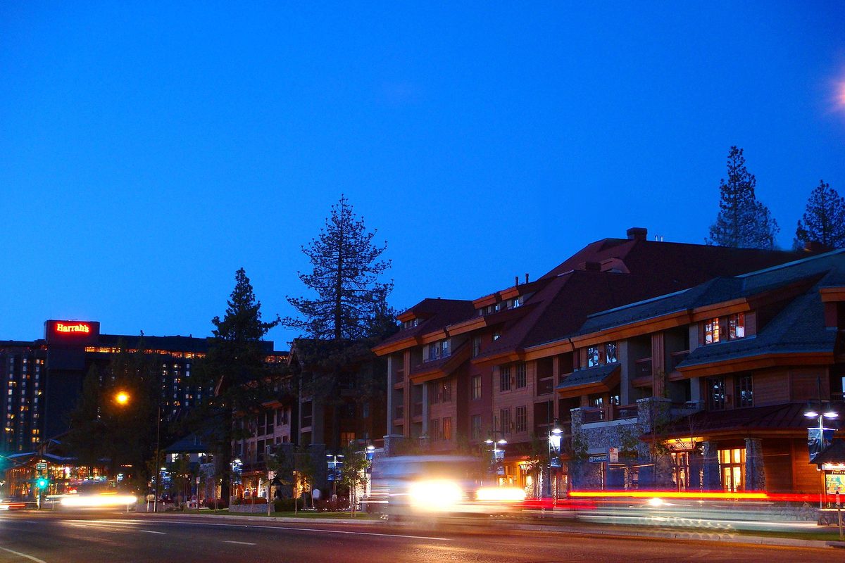 Lodge-style hotel buildings and tall pines along a South Lake Tahoe street at dusk, with headlights streaking past