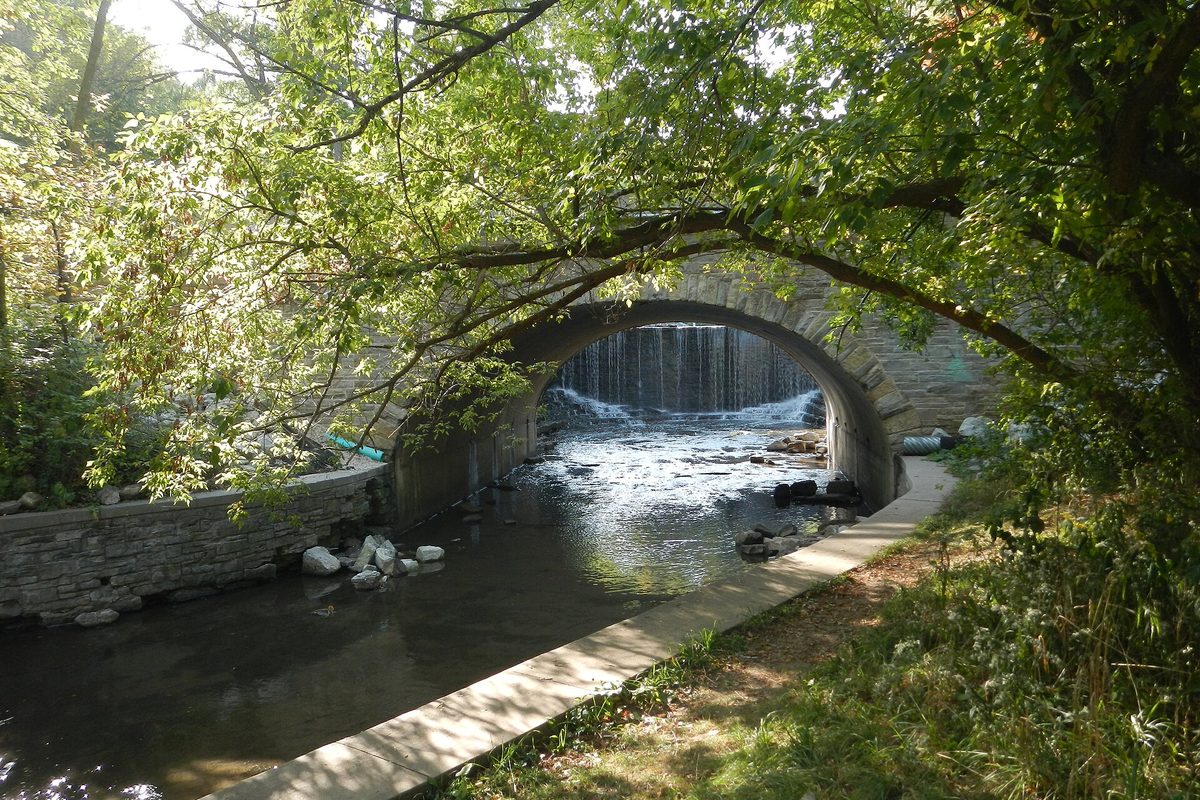 A stone arch bridge over a small waterfall on Oak Creek, framed by overhanging trees, with a concrete path along the stream