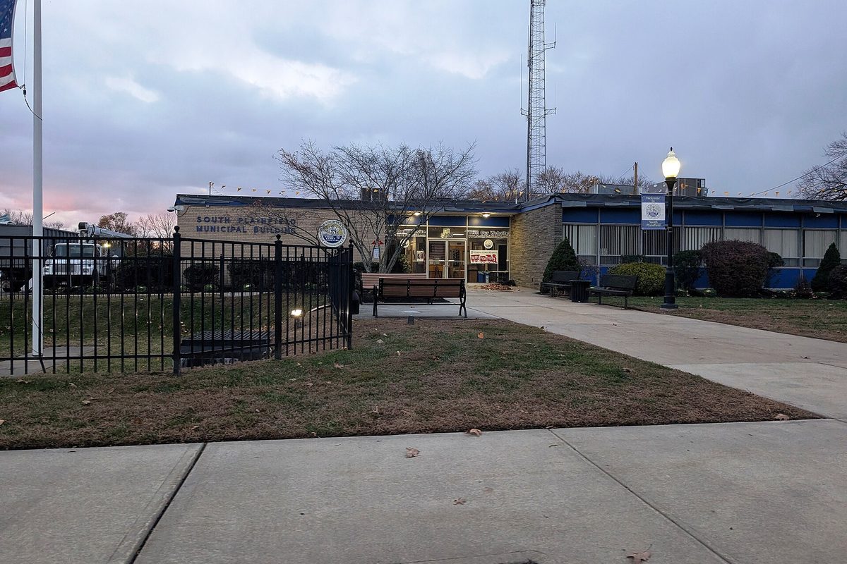 The low brick and blue-panel South Plainfield Municipal Building behind a black iron fence at dusk, with a flagpole and a radio tower