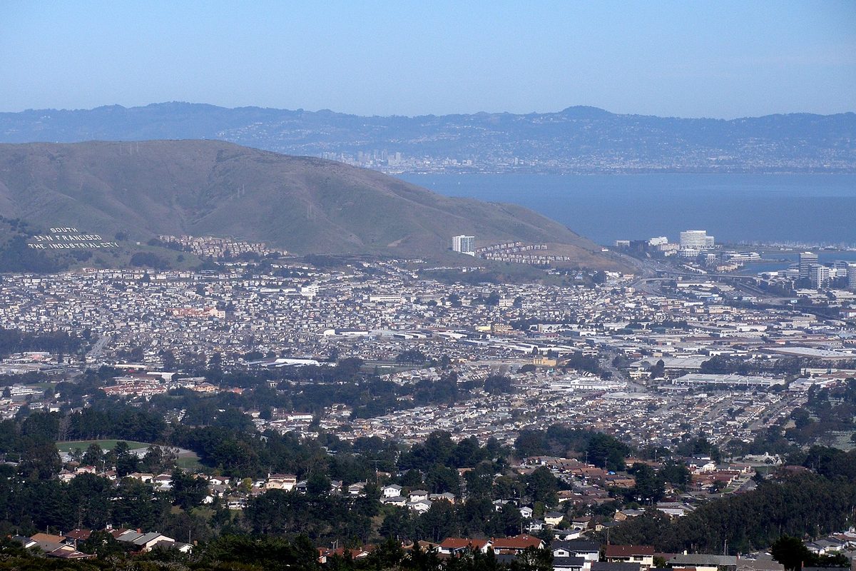 South San Francisco seen from Sweeney Ridge: rows of houses filling the valley, a hillside with the city's name in white letters, and the bay behind