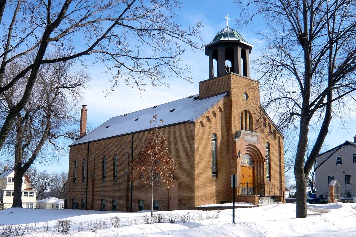 The tan brick St. Sava Serbian Orthodox Church in South St. Paul with a copper-domed bell tower, snow on the roof and lawn, under a blue sky