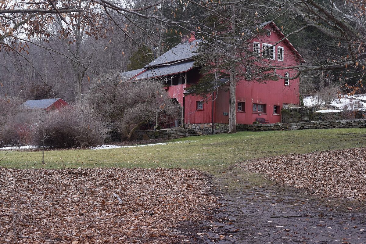 A red barn-style building with a gray roof behind leafless shrubs, with fallen leaves and patches of snow on the lawn at the Audubon Center at Bent of the River in Southbury
