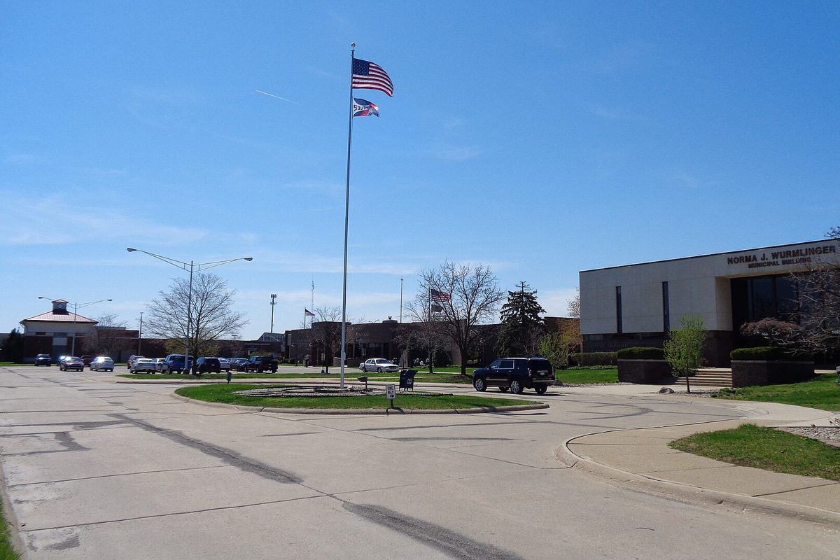 Southgate Municipal Complex, with American and Michigan flags on a tall pole over a traffic circle, a parking lot and the Norma J. Wurmlinger Memorial Building under a clear blue sky