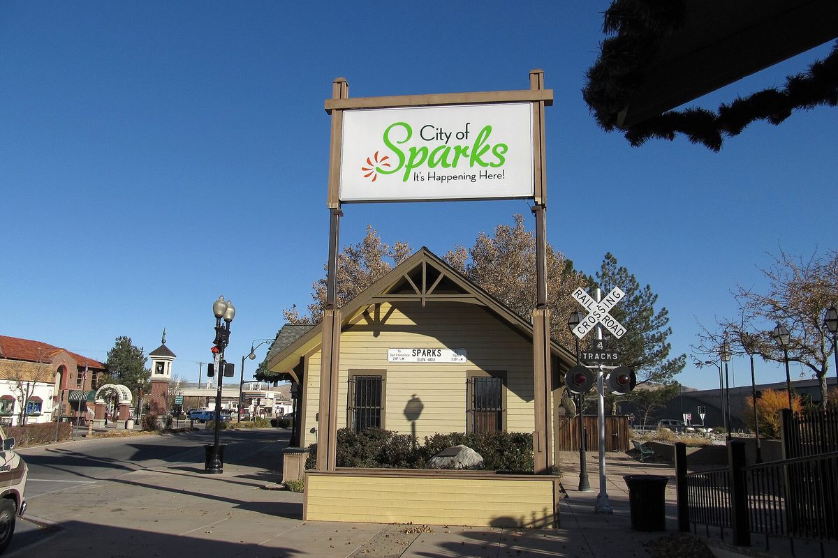 A City of Sparks sign above a small yellow wooden depot building beside a railroad crossing sign in Victorian Square, under a deep blue sky