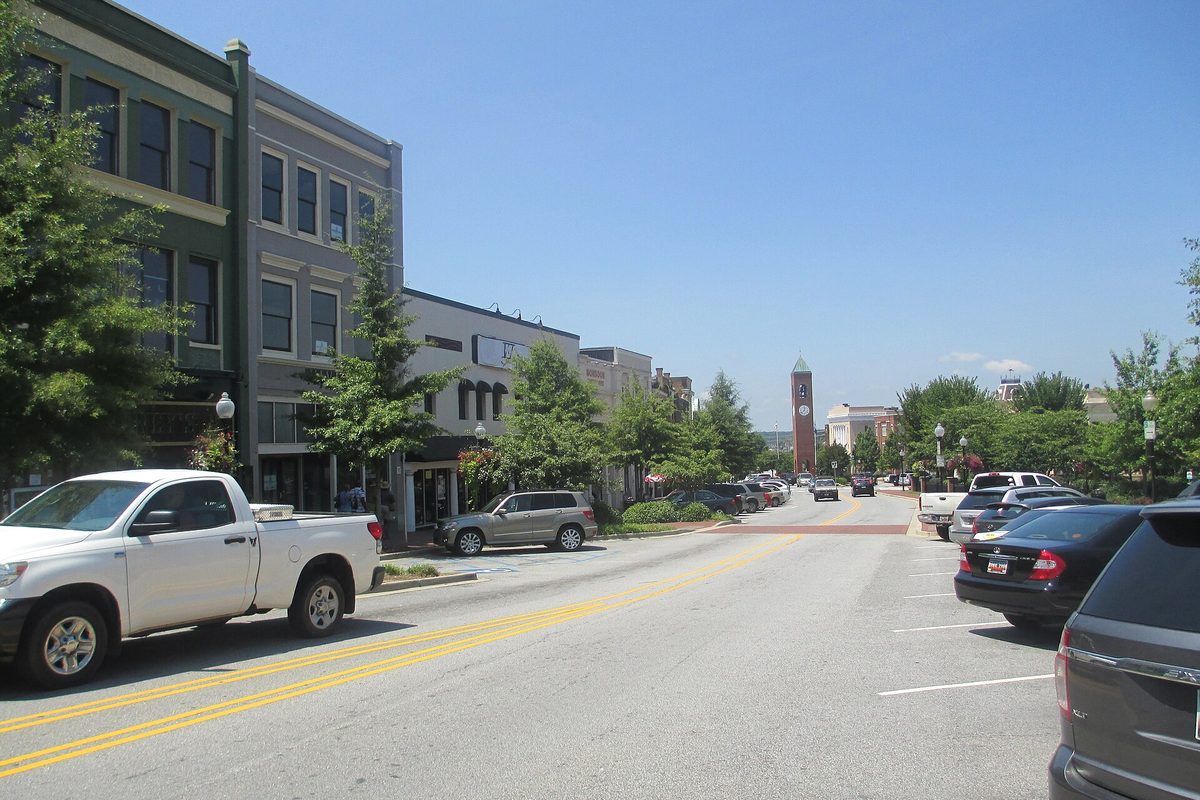 A downtown Spartanburg street with a white pickup and parked cars beside two- and three-story buildings, and a brick clock tower at the far end