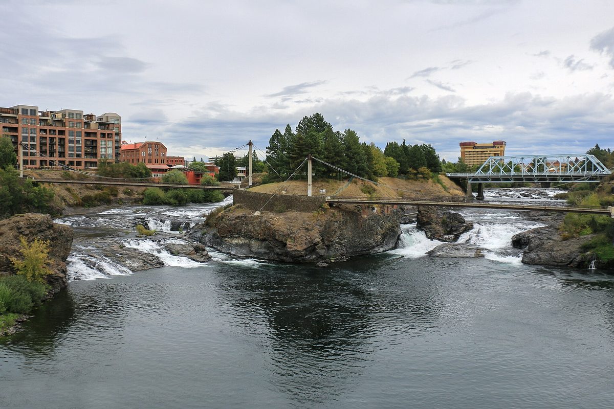 Water pouring over dark basalt at the Upper Spokane Falls, with a cable footbridge, a steel truss bridge and downtown buildings behind