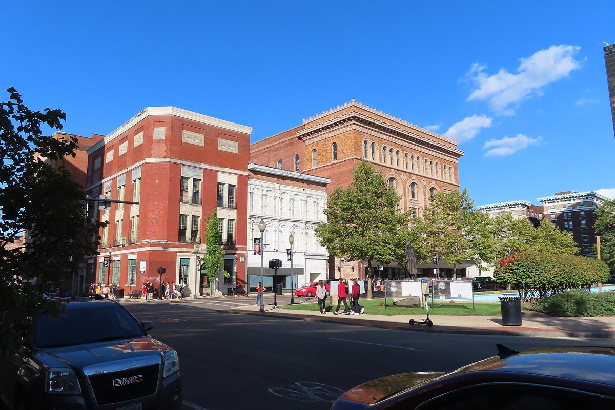 Red brick and ornate stone buildings on a sunny downtown Springfield corner, with people walking along the sidewalks and the hood of a parked SUV in the foreground
