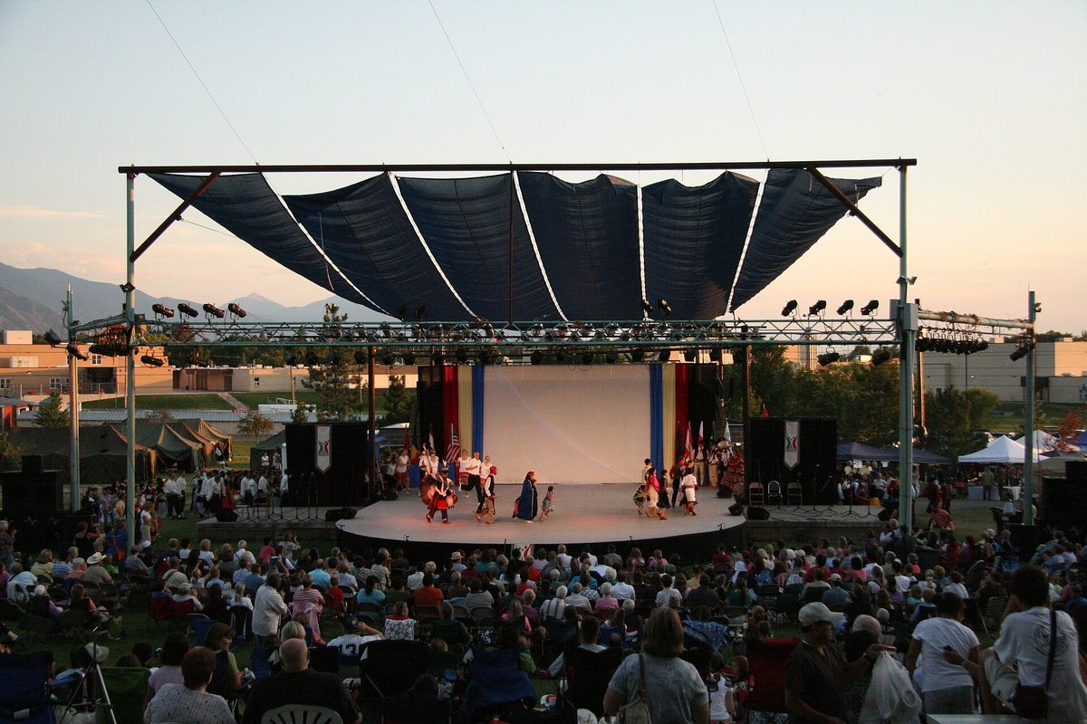 Dancers in bright costumes on an outdoor stage under a dark canopy at Springville's World Folkfest, with a large crowd seated on the lawn at dusk
