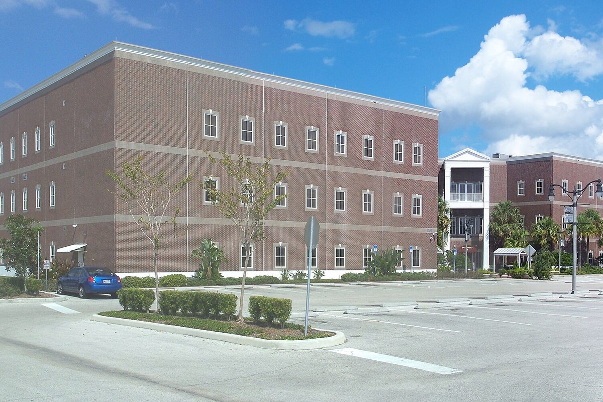 St. Cloud's three-story brick city hall beside a parking lot with a blue car, young trees and a covered entrance at right