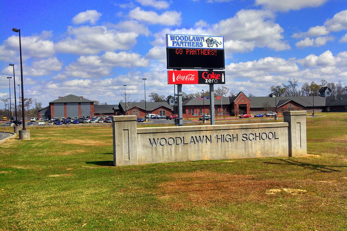 The stone Woodlawn High School entrance sign and a Woodlawn Panthers marquee on a lawn, with the brick school buildings and a full parking lot behind them under a blue sky