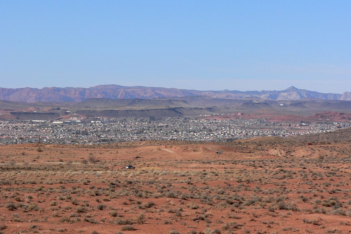 St. George spread across a wide valley below layered red-rock ridges, seen from red desert scrubland east of town