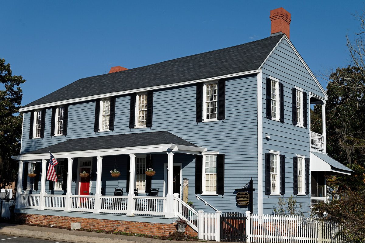 A two-story blue clapboard historic house in St. Marys with black shutters, a white front porch, a red door, an American flag and a white picket fence under a clear blue sky