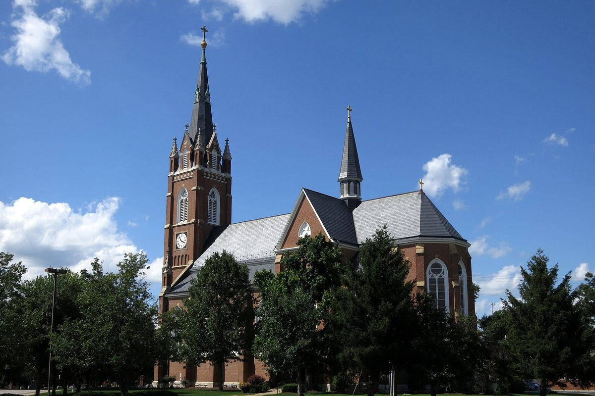 All Saints Catholic Church in St. Peters, a red-brick church with a clock tower and a second slate spire, behind evergreens under a blue sky