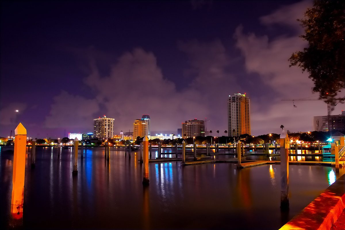 Downtown St. Petersburg high-rises lit up at night, their lights reflected in the still water of a marina lined with wooden pilings