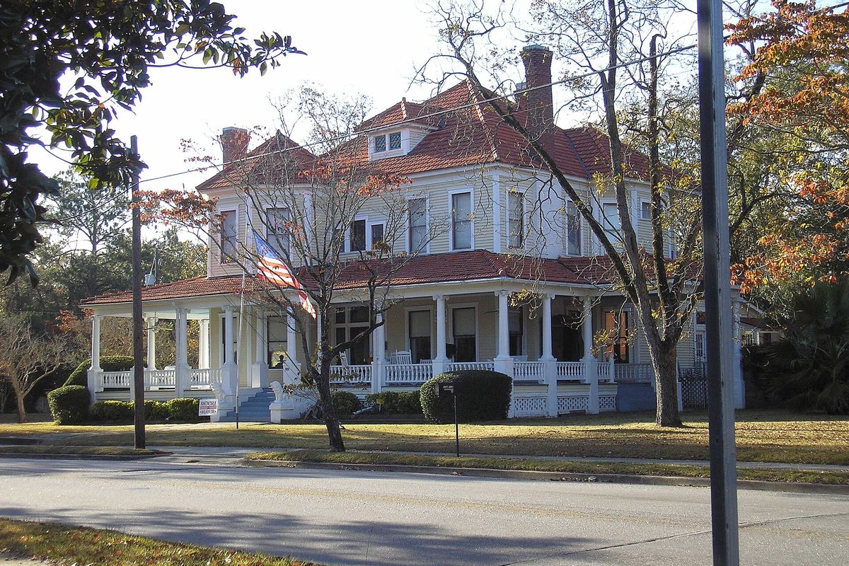 A large white two-story house with a red tile roof, dormers and a wraparound porch behind bare and autumn trees in Statesboro's Savannah Avenue Historic District
