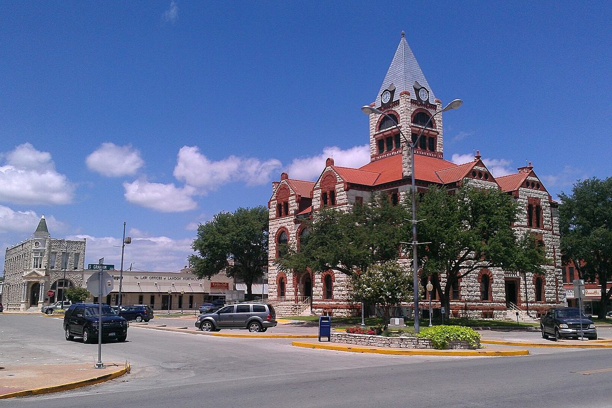 The red-roofed stone Erath County Courthouse with its pointed clock tower, seen across a street corner in Stephenville under a blue sky with white clouds