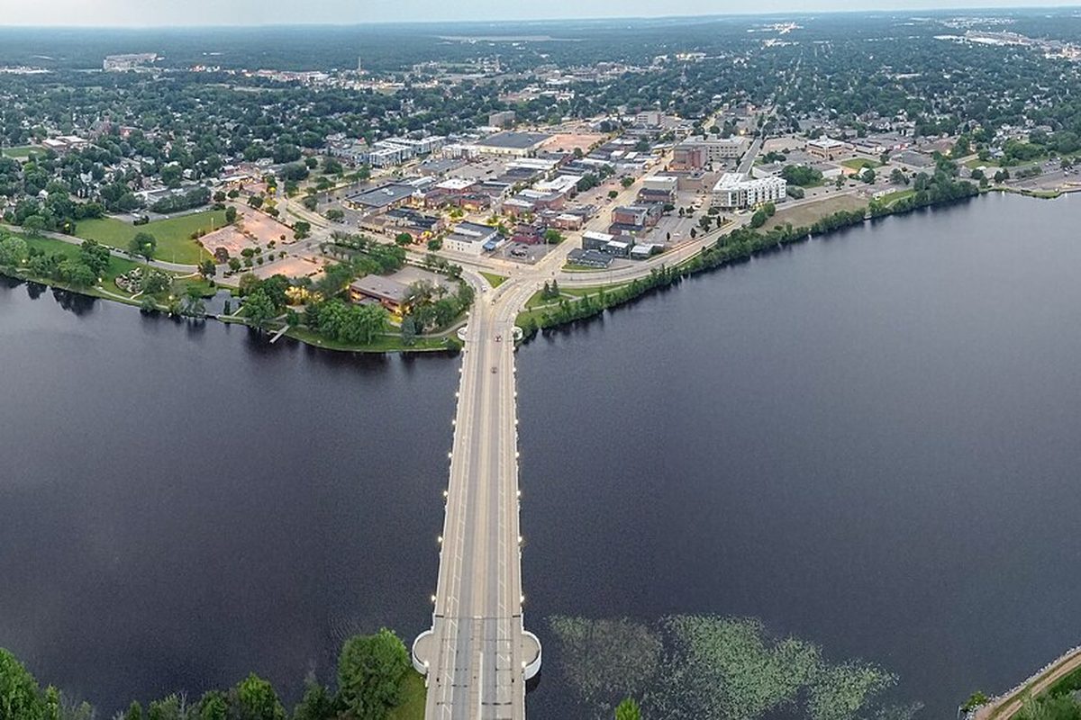 Aerial view of a long bridge crossing a wide river into downtown Stevens Point, with low buildings and trees on the far bank