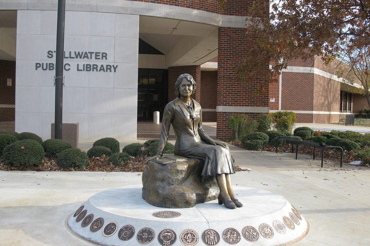A bronze statue of historian Angie Debo seated on a rock outside the brick and stone Stillwater Public Library, on a round base ringed with medallions