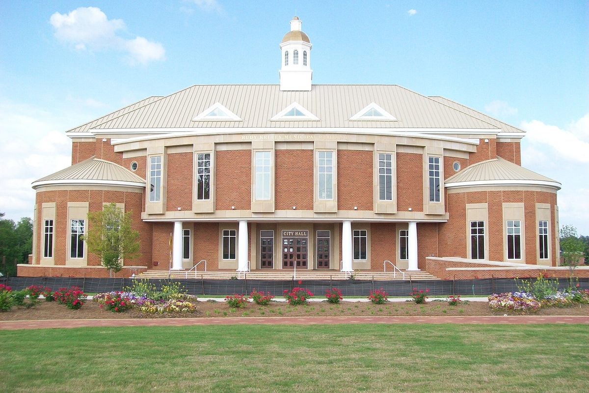 Stockbridge City Hall, a red brick building with white columns and a white cupola, behind flower beds and a mown lawn under a blue sky