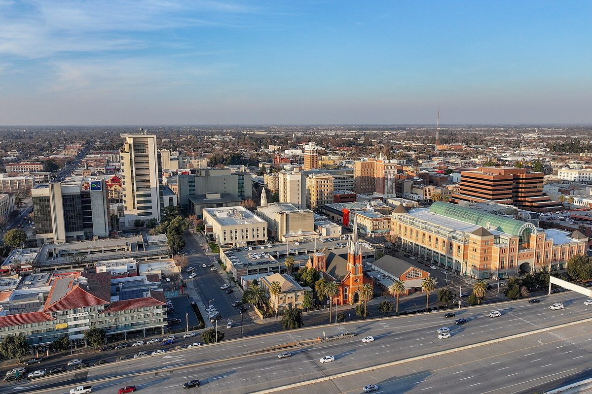 Aerial view of downtown Stockton in late-afternoon light, with a red brick church spire, mid-rise towers, palm trees and a freeway in the foreground