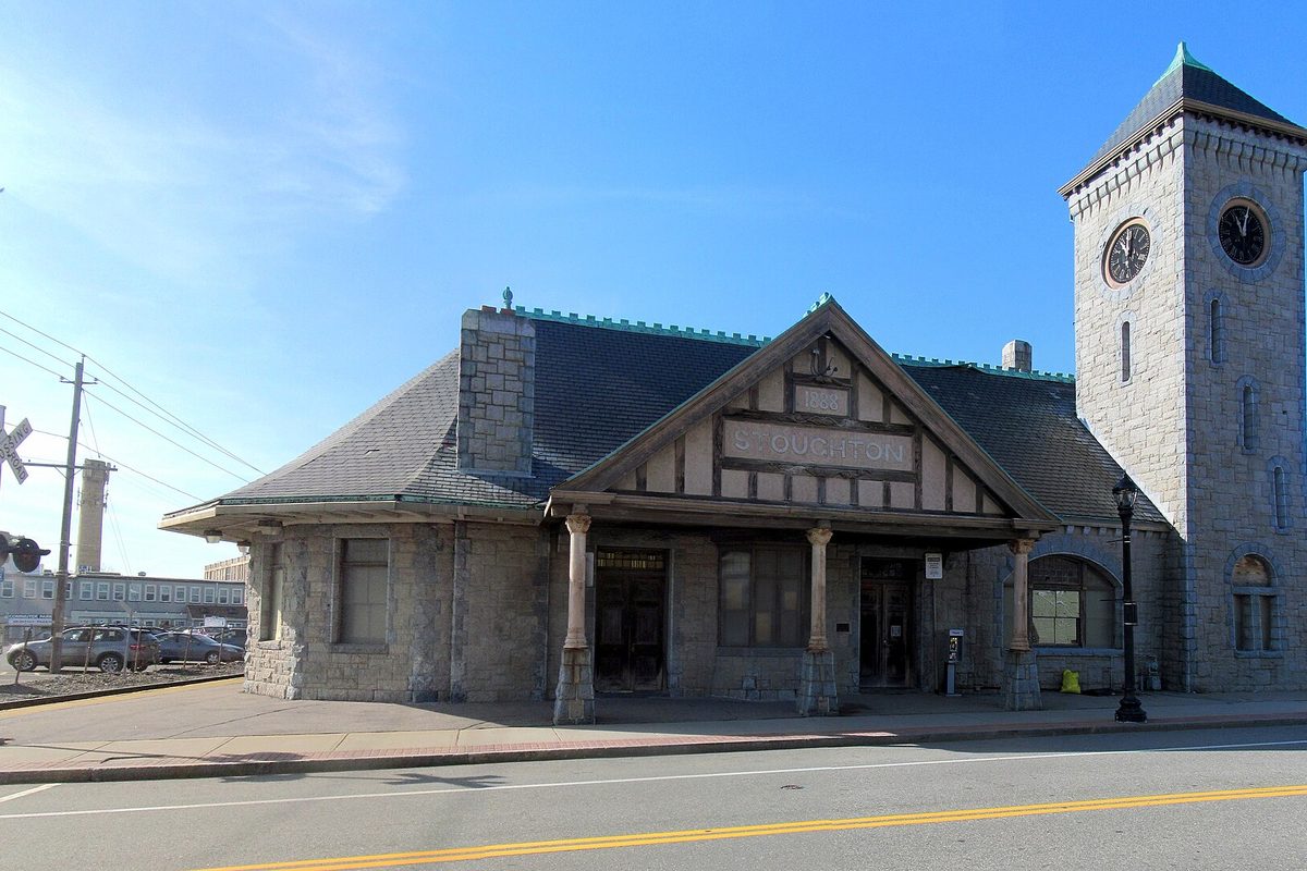 The granite Stoughton train station with its clock tower and half-timbered gable, seen across Wyman Street on a clear day
