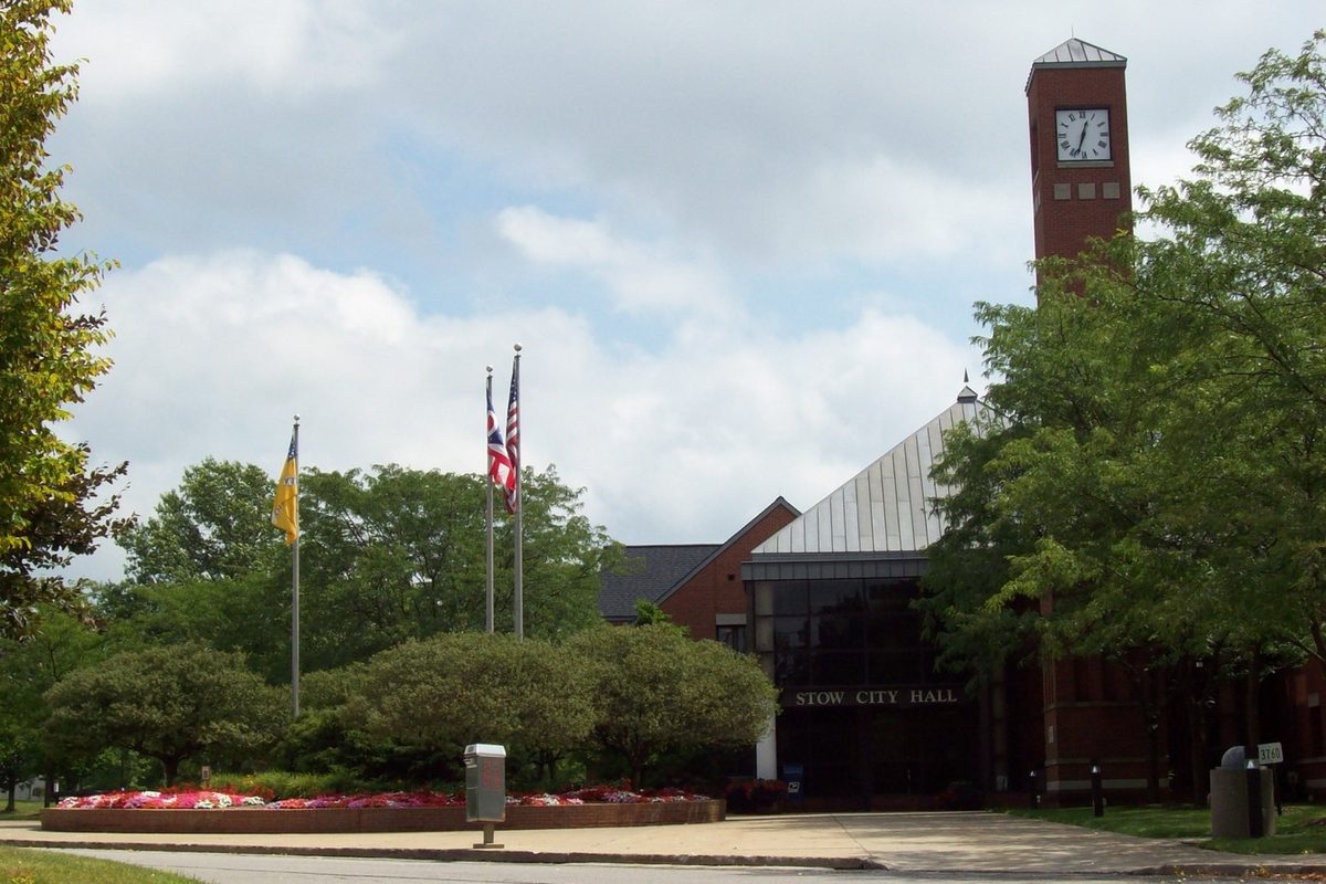 Stow City Hall, a brick building with a square clock tower, seen past flagpoles, a red flower bed and shade trees